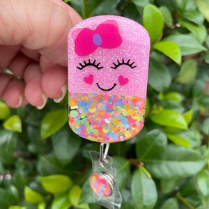 May include: Pink glitter badge holder with a smiling face, a pink bow, and confetti sprinkles. The holder is attached to a clear retractable badge reel.