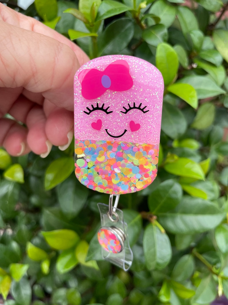 May include: Pink glitter badge holder with a smiling face, a pink bow, and confetti sprinkles. The holder is attached to a clear retractable badge reel.