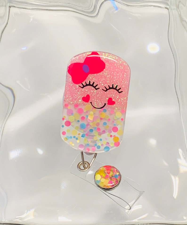 May include: A pink and white glitter badge holder with a smiling face, pink bow, and confetti design. The holder has a clear plastic retractable badge reel with a colourful confetti design.