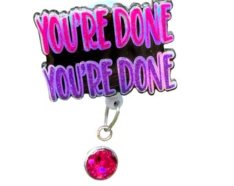 May include: A badge reel with the text "YOU'RE DONE" in pink and purple glitter lettering. The badge reel has a black background and a small, round, pink glitter charm attached to the bottom. The reel is silver.