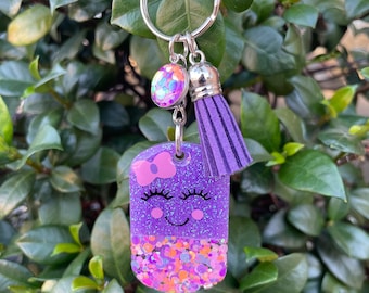 May include: Purple glitter keychain with a smiling face, eyelashes, and a pink bow. The keychain has a purple tassel and a clear iridescent charm.