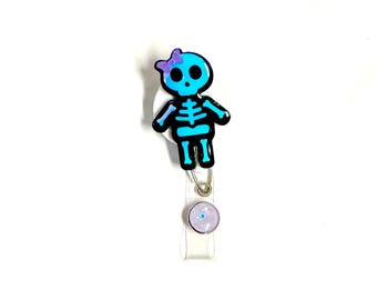 May include: A blue and black skeleton-shaped badge reel with a purple bow. The badge reel has a clear plastic strap and a round, iridescent button. The skeleton design is detailed with a black outline and a blue body.