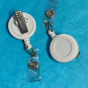 May include: Two white retractable badge holders with clear plastic clips. The holders have a silver clip and a silver ring.