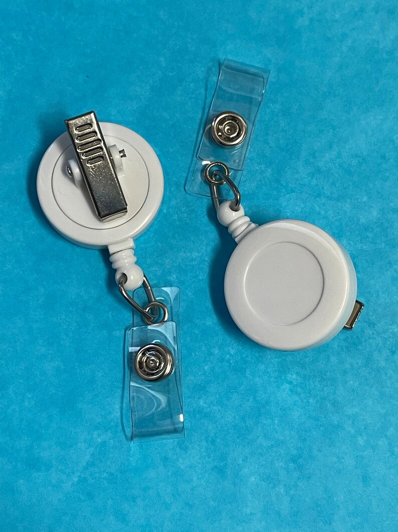 May include: Two white retractable badge holders with clear plastic clips. The holders have a silver clip and a silver ring.