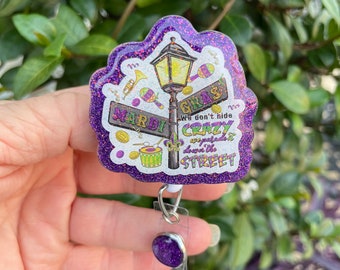 May include: A purple glitter badge reel with a Mardi Gras theme. The design includes a street lamp, musical instruments, and the words "Mardi Gras" and "We don't hide crazy, we parade it down the street."