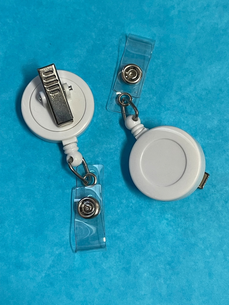May include: Two white retractable badge holders with clear plastic clips. The holders have a silver clip and a silver ring.