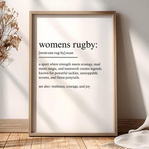 Könnte beinhalten: Ein gerahmter Druck mit dem Text "womens rugby:" und einer Definition. Die Definition beschreibt Rugby als Sport, in dem Stärke auf Strategie trifft, Teamwork Legenden erschafft und wilde Pferdeschwänze üblich sind. Der Rahmen ist hellbraun.