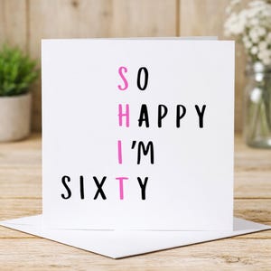 May include: A square white greeting card with black text that reads SO HAPPY I'M SIXTY. The letters S, H, I, and T are arranged vertically in a column and are colored in bright pink, forming a word that stands out against the black text.