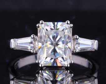 Può includere: Un anello d'argento con una grande pietra centrale rettangolare in moissanite trasparente e due pietre in moissanite più piccole, a forma di baguette, su entrambi i lati.