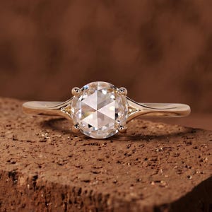 May include: A gold ring featuring a round rose-cut center stone held by four prongs. The band is smooth and tapers toward the setting, which includes small triangular cutouts on each side of the stone, creating a clean and polished appearance.