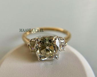 Puede incluir: Un anillo de oro con un gran diamante de talla cojín en el centro y dos diamantes más pequeños a cada lado. El anillo está sobre una superficie blanca.