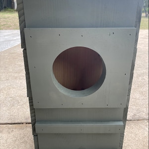 May include: A gray wooden birdhouse with a round entrance hole. The birdhouse has a simple design with a flat roof and a few horizontal boards on the front.