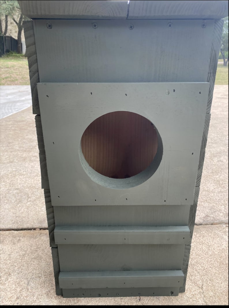 May include: A gray wooden birdhouse with a round entrance hole. The birdhouse has a simple design with a flat roof and a few horizontal boards on the front.