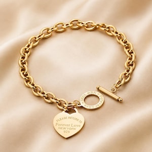 Könnte beinhalten: Ein goldfarbenes Gliederkettarmband mit Knebelverschluss und herzförmigem Anhänger. Der Anhänger ist mit dem Text PLEASE RETURN TO Forever Love NEW YORK 925 graviert, während der kreisförmige Knebelring die Gravur Forever Love NEW YORK aufweist.