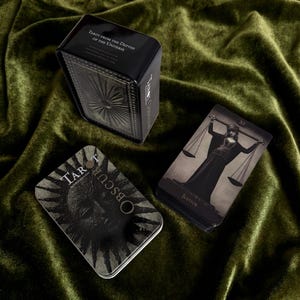 May include: A black tarot card deck with a matching tin box, and a separate tin with the word "OBSCUR" printed on it. The tarot cards feature illustrations, and the box has the text "TAROT FROM THE DEPTHS OF THE UNIVERSE". The background is a dark green velvet.