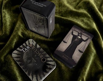 Op de afbeelding: Een zwarte metalen doos met het label Tarot from the Depths of the Universe, een rechthoekig blik met een zongrafiek met het label Tarot Obscura, en een tarotkaart met een figuur die een weegschaal vasthoudt met het label Justice en Romeins cijfer XI.