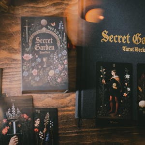 May include: A Secret Garden tarot deck box with floral illustrations and a matching black guidebook featuring gold lettering. Several tarot cards are displayed, including the Queen, Ace of Swords, and Ace of Cups, all featuring dark backgrounds.