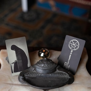 May include: A dark metal inkwell with a rounded lid sits between two rectangular cards. The left card features a black and white illustration of a hooded figure, while the right card displays a detailed graphic of a skeleton key with a circular head.