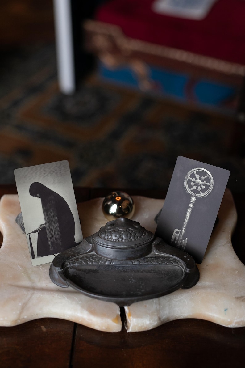 May include: A dark metal inkwell with a rounded lid sits between two rectangular cards. The left card features a black and white illustration of a hooded figure, while the right card displays a detailed graphic of a skeleton key with a circular head.
