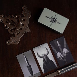 May include: A deck of tarot cards with a light green back featuring a star design, three cards showing a sword, a horned figure, and hands holding a sword, a metal protractor, and a letter opener with a dark handle and silver metal blade.