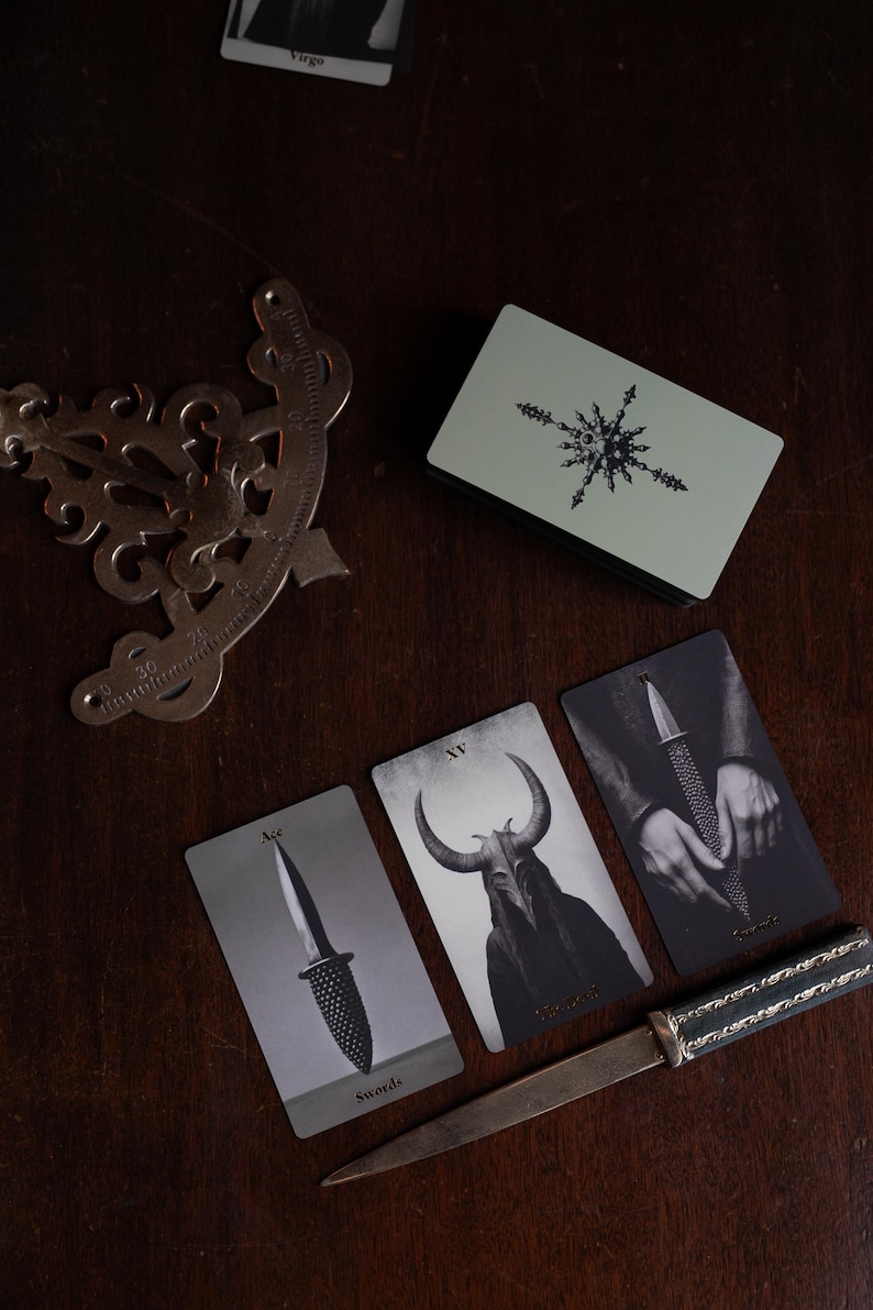 May include: A deck of tarot cards with a light green back featuring a star design, three cards showing a sword, a horned figure, and hands holding a sword, a metal protractor, and a letter opener with a dark handle and silver metal blade.