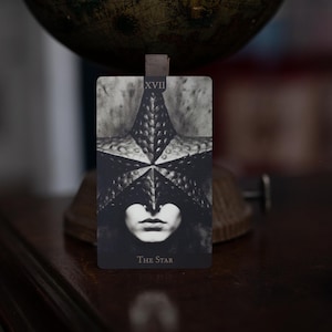 May include: A rectangular tarot card titled The Star, labeled XVII, featuring a black and white illustration of a person wearing a textured, star-shaped mask over their eyes. The card displays a high-contrast, monochromatic design with a dark background.
