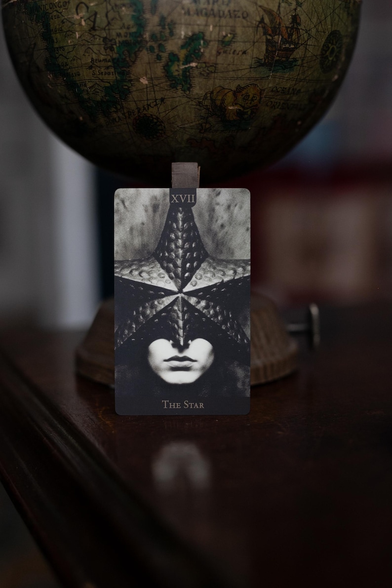 May include: A rectangular tarot card titled The Star, labeled XVII, featuring a black and white illustration of a person wearing a textured, star-shaped mask over their eyes. The card displays a high-contrast, monochromatic design with a dark background.