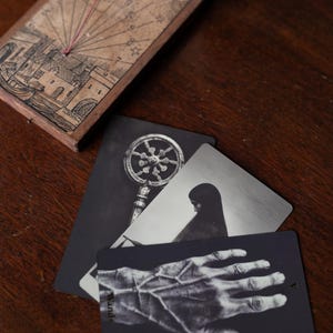 May include: A wooden sundial with a string gnomon and engraved illustrations of a town and celestial markings, accompanied by three rectangular cards featuring black and white imagery of a key, a hooded figure, and a hand with prominent veins.
