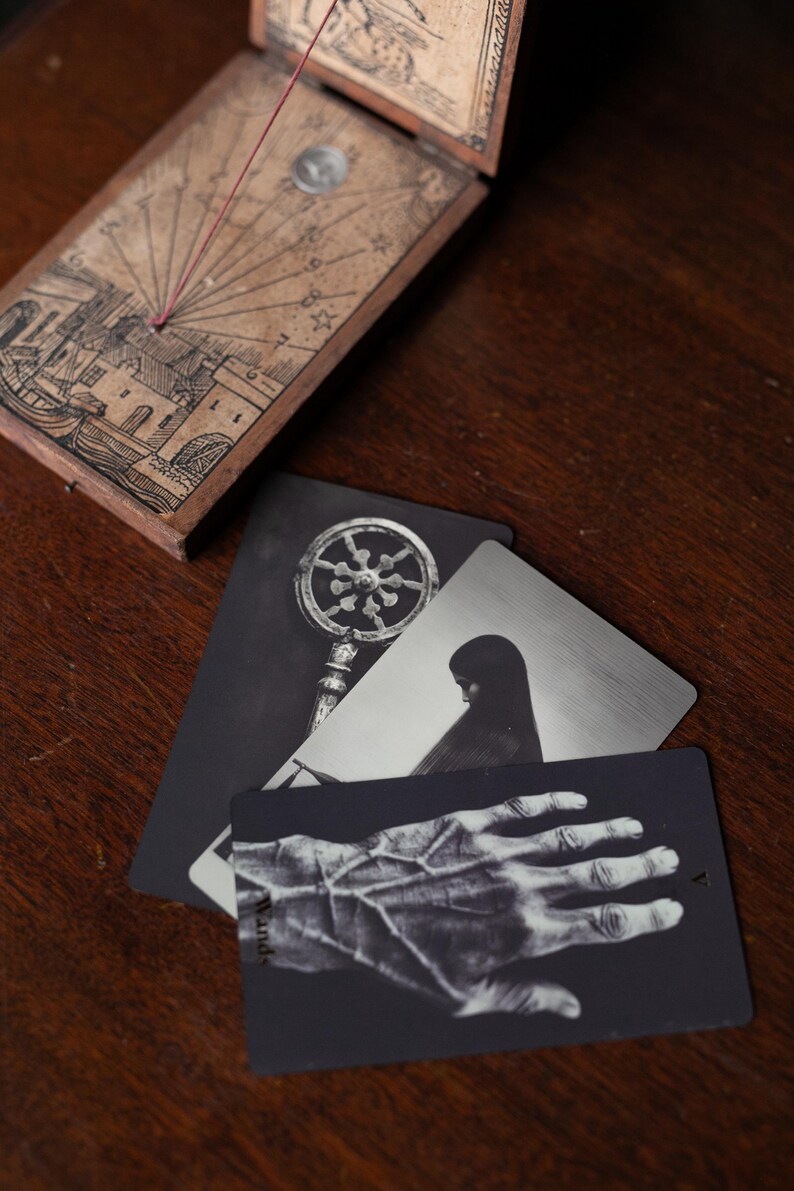 May include: A wooden sundial with a string gnomon and engraved illustrations of a town and celestial markings, accompanied by three rectangular cards featuring black and white imagery of a key, a hooded figure, and a hand with prominent veins.