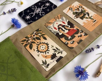 Op de afbeelding: Een groene stoffen tarotkaarthouder met drie uitgestalde kaarten: Aas van Pentakels, Gematigdheid en de 9 van Pentakels. Een zwarte kaart met witte ster- en oogmotieven ligt ernaast. De houder is versierd met groene ster- en lijnpatronen.