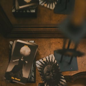May include: A deck of tarot cards with black and white imagery. One card shows a figure with a moon head labeled XXI The World, and another card displays a sun face labeled XIX The Sun. The cards feature gold text and are arranged on a surface.