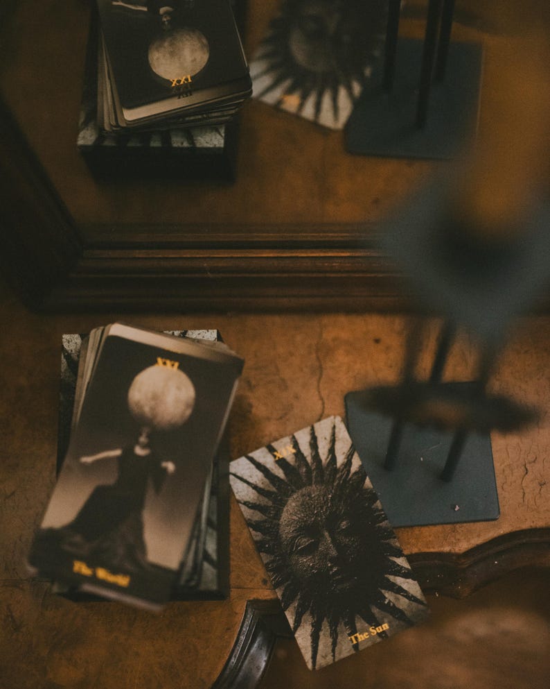 May include: A deck of tarot cards with black and white imagery. One card shows a figure with a moon head labeled XXI The World, and another card displays a sun face labeled XIX The Sun. The cards feature gold text and are arranged on a surface.