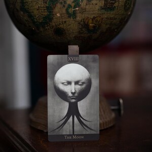 May include: A rectangular tarot card titled The Moon with the Roman numeral XVIII at the top. The card features a monochromatic, surreal illustration of a pale, spherical head with closed eyes, supported by dark, tentacle-like structures at the base.