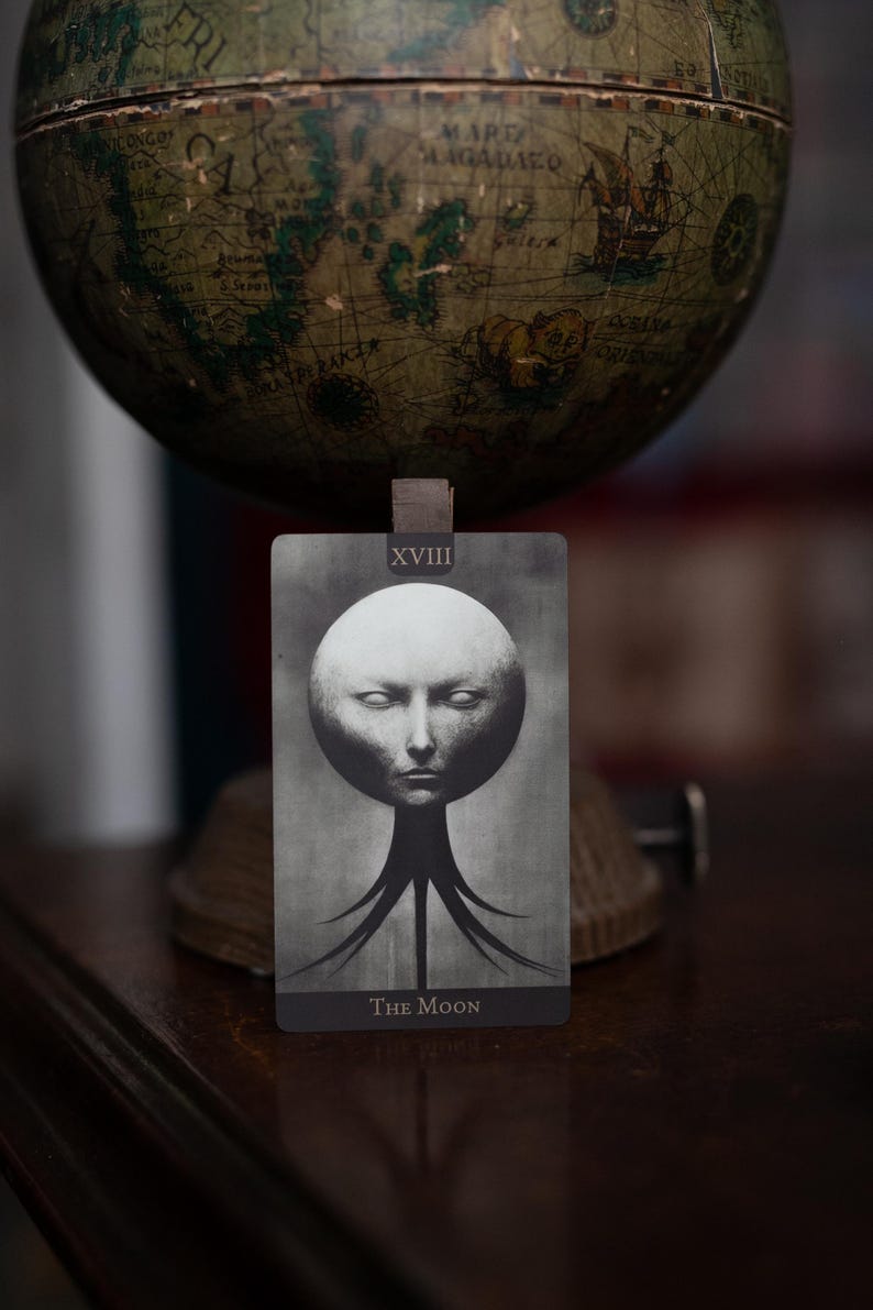 May include: A rectangular tarot card titled The Moon with the Roman numeral XVIII at the top. The card features a monochromatic, surreal illustration of a pale, spherical head with closed eyes, supported by dark, tentacle-like structures at the base.