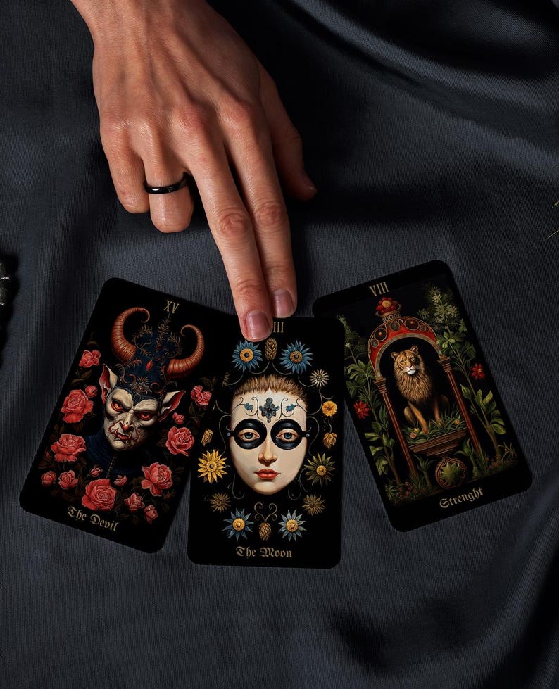 May include: Three tarot cards with detailed illustrations are displayed. The cards feature images of The Devil, The Moon, and Strength. The Devil card shows a horned figure surrounded by roses. The Moon card has a face with floral accents. The Strength card depicts a lion.