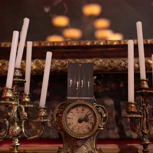 May include: A tarot card labeled IV Wands featuring four lit candles stands atop an ornate, aged brass clock. The clock is flanked by two matching brass candelabras, each holding multiple thin, white, unlit candles with visible wicks.