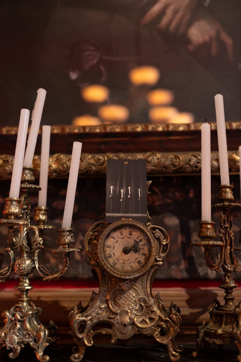 May include: A tarot card labeled IV Wands featuring four lit candles stands atop an ornate, aged brass clock. The clock is flanked by two matching brass candelabras, each holding multiple thin, white, unlit candles with visible wicks.