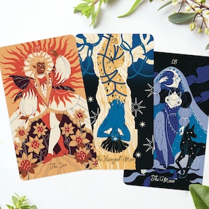 Op de afbeelding: Drie tarotkaarten met kleurrijke illustraties. De Zon-kaart toont een figuur in een vurige outfit, de Hanged Man-kaart toont een blauwe figuur ondersteboven en de Maan-kaart toont een figuur met een zwarte hond.