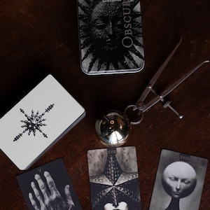 May include: A Tarot Obscura deck in a metal tin, a deck of cards with a star-like symbol, a metallic sphere, and a metal caliper. Three tarot cards are displayed: V Wands featuring a hand, XVII The Star, and XVIII The Moon with a surreal head.