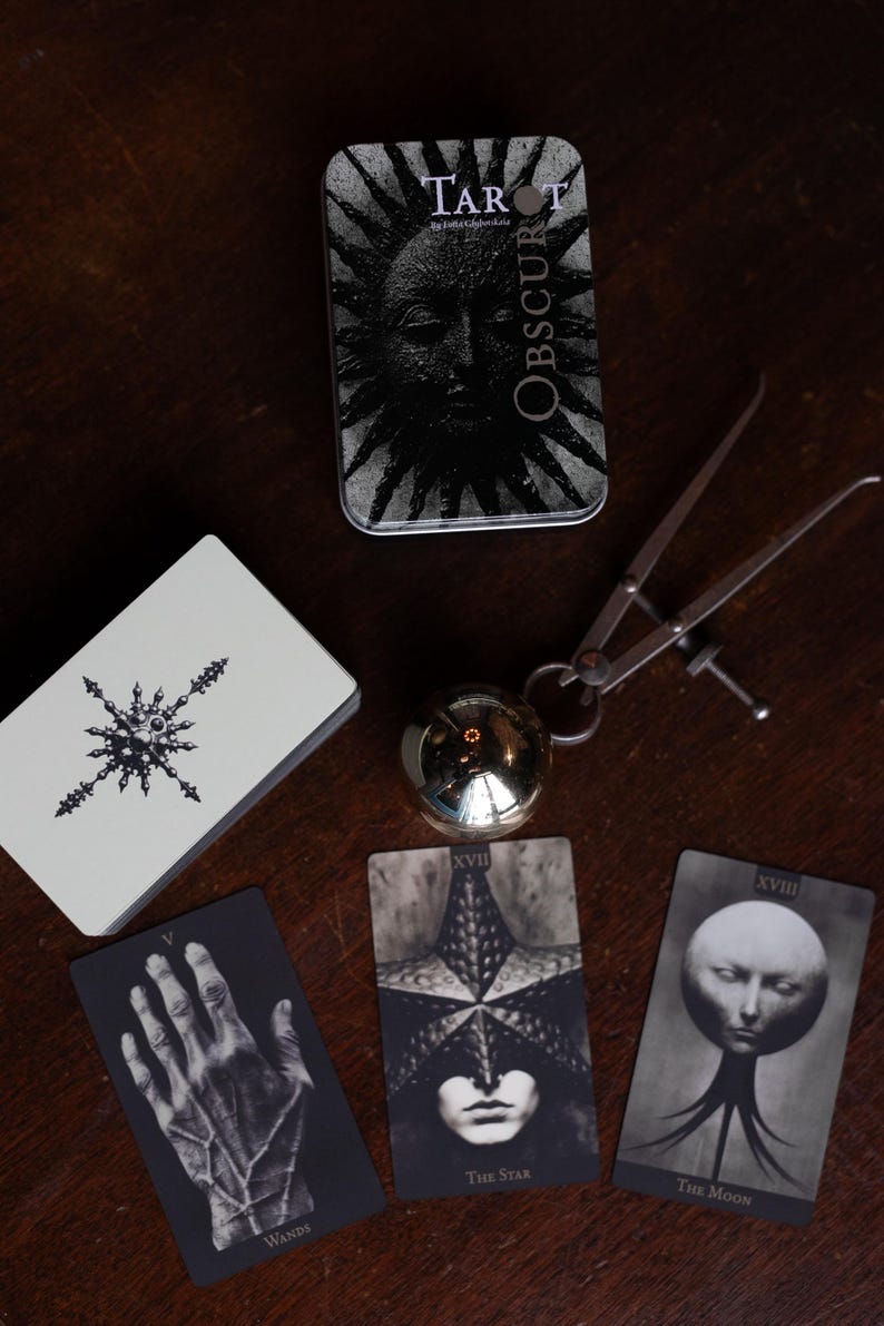 May include: A Tarot Obscura deck in a metal tin, a deck of cards with a star-like symbol, a metallic sphere, and a metal caliper. Three tarot cards are displayed: V Wands featuring a hand, XVII The Star, and XVIII The Moon with a surreal head.