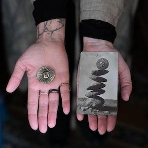 May include: A round brass-colored metal coin featuring a raised sun face design, held alongside a rectangular tarot card labeled Ace of Pentacles, which depicts a stack of stones topped with a matching sun face icon in a monochromatic gray tone.