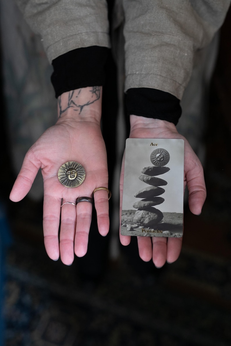 May include: A round brass-colored metal coin featuring a raised sun face design, held alongside a rectangular tarot card labeled Ace of Pentacles, which depicts a stack of stones topped with a matching sun face icon in a monochromatic gray tone.