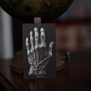 May include: A rectangular tarot card with a black background featuring a detailed white illustration of a human hand with prominent veins. The Roman numeral V is printed at the top center, and the word WANDS is printed at the bottom center.
