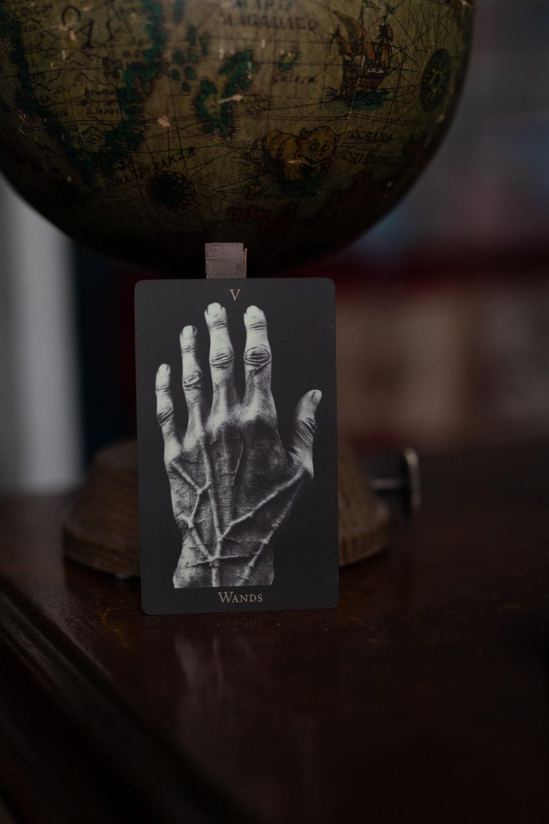 May include: A rectangular tarot card with a black background featuring a detailed white illustration of a human hand with prominent veins. The Roman numeral V is printed at the top center, and the word WANDS is printed at the bottom center.