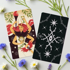 Op de afbeelding: Twee tarotkaarten met illustraties. E&eacute;n kaart toont een vrouw met rood haar, die een fakkel en een kelk vasthoudt. De andere kaart heeft een zwarte achtergrond met witte sterren en geometrische ontwerpen. Bloemenaccenten omringen de kaarten.