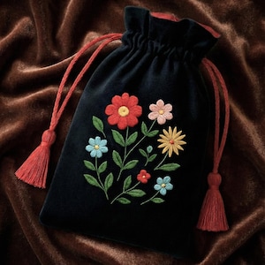 Puede incluir: Una bolsa de terciopelo negro con cordón y diseño floral bordado. Las flores son rojas, rosas, amarillas y azules, con hojas verdes. La bolsa tiene cordones y borlas rojas.