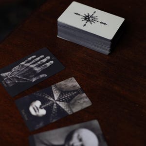May include: A deck of rectangular cards with a black and white star-like emblem on the back, accompanied by three face-up cards featuring monochromatic illustrations of a hand, a star-shaped figure, and a circular form on a dark surface.