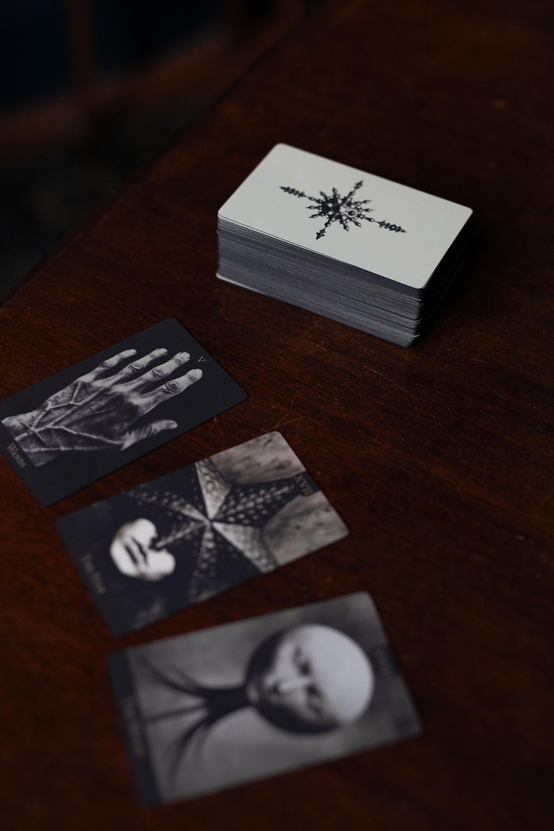 May include: A deck of rectangular cards with a black and white star-like emblem on the back, accompanied by three face-up cards featuring monochromatic illustrations of a hand, a star-shaped figure, and a circular form on a dark surface.