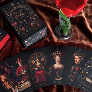 May include: A black tarot deck box and cards with floral and botanical illustrations. The box reads "Secret Garden Tarot Deck". The cards feature portraits of figures with floral accents. A red flower-shaped candle holder is also present.