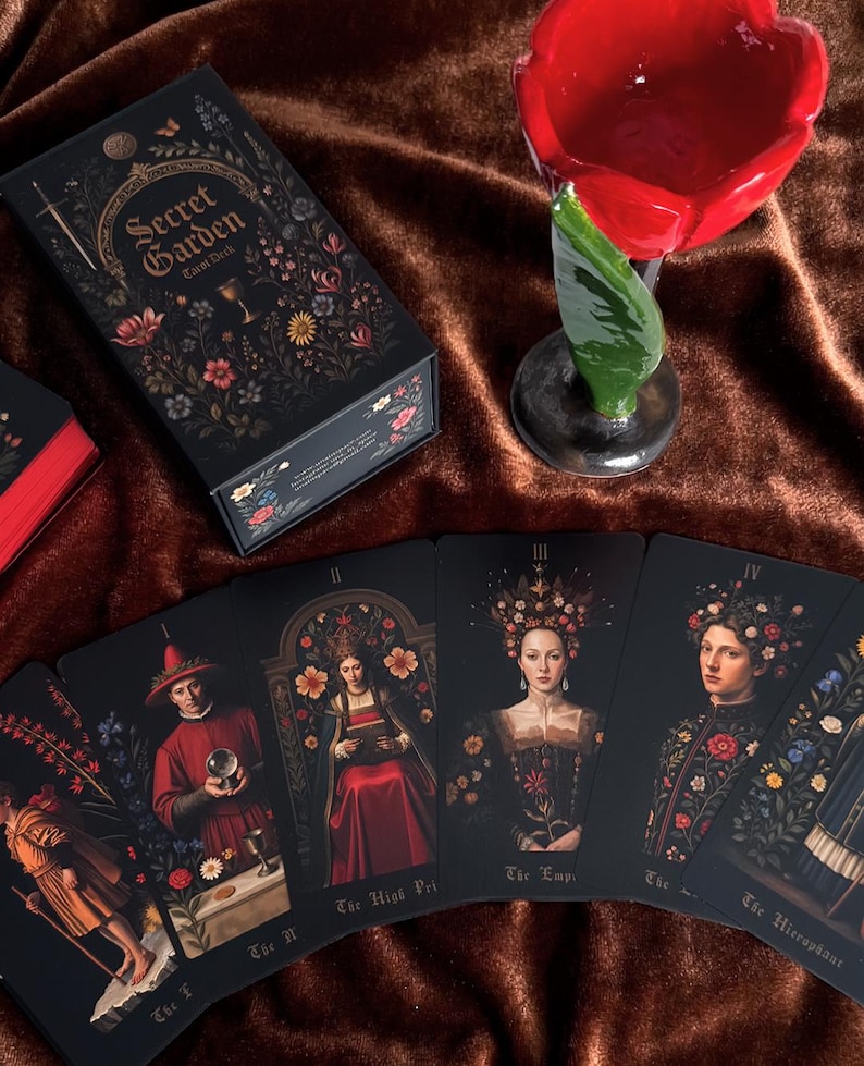 May include: A black tarot deck box and cards with floral and botanical illustrations. The box reads "Secret Garden Tarot Deck". The cards feature portraits of figures with floral accents. A red flower-shaped candle holder is also present.